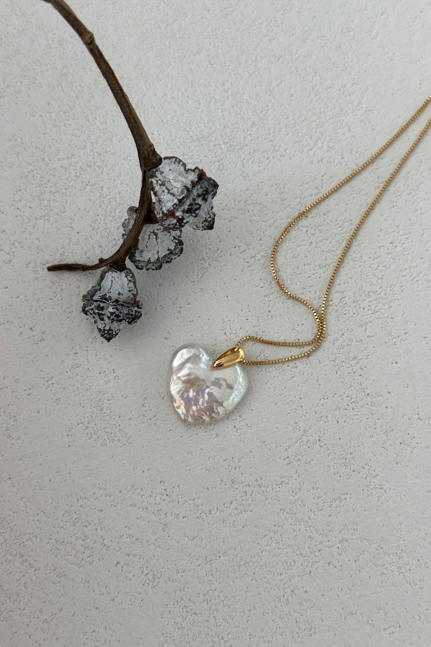 The Large Heart Pendant Necklace by Siren Mode, with an 18K gold-plated sterling silver chain and a 14K gold clasp, is elegantly displayed against a textured white surface. The necklace features a shimmering iridescent heart-shaped pendant and its chain, arranged in gentle loops, showcases both gold and sterling silver elements. A small dried branch with dark, wilted flowers rests beside it, adding a contrasting element.