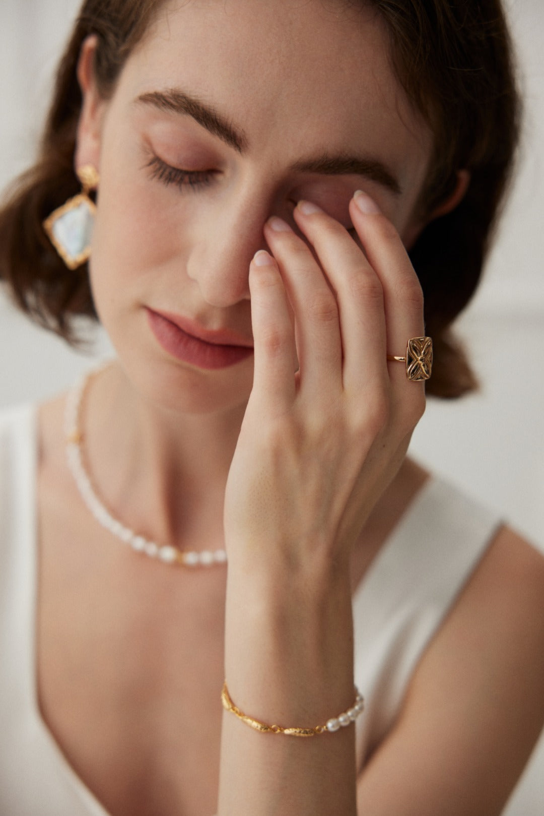 A woman with short hair is gently touching her face with her eyes closed. She is wearing a gold ring, the Lucky Clover Adjustable Ring in Vintage Gold Finish by Siren Mode, and a pearl necklace. A bracelet and large earrings complement her white sleeveless top. She appears calm and serene.