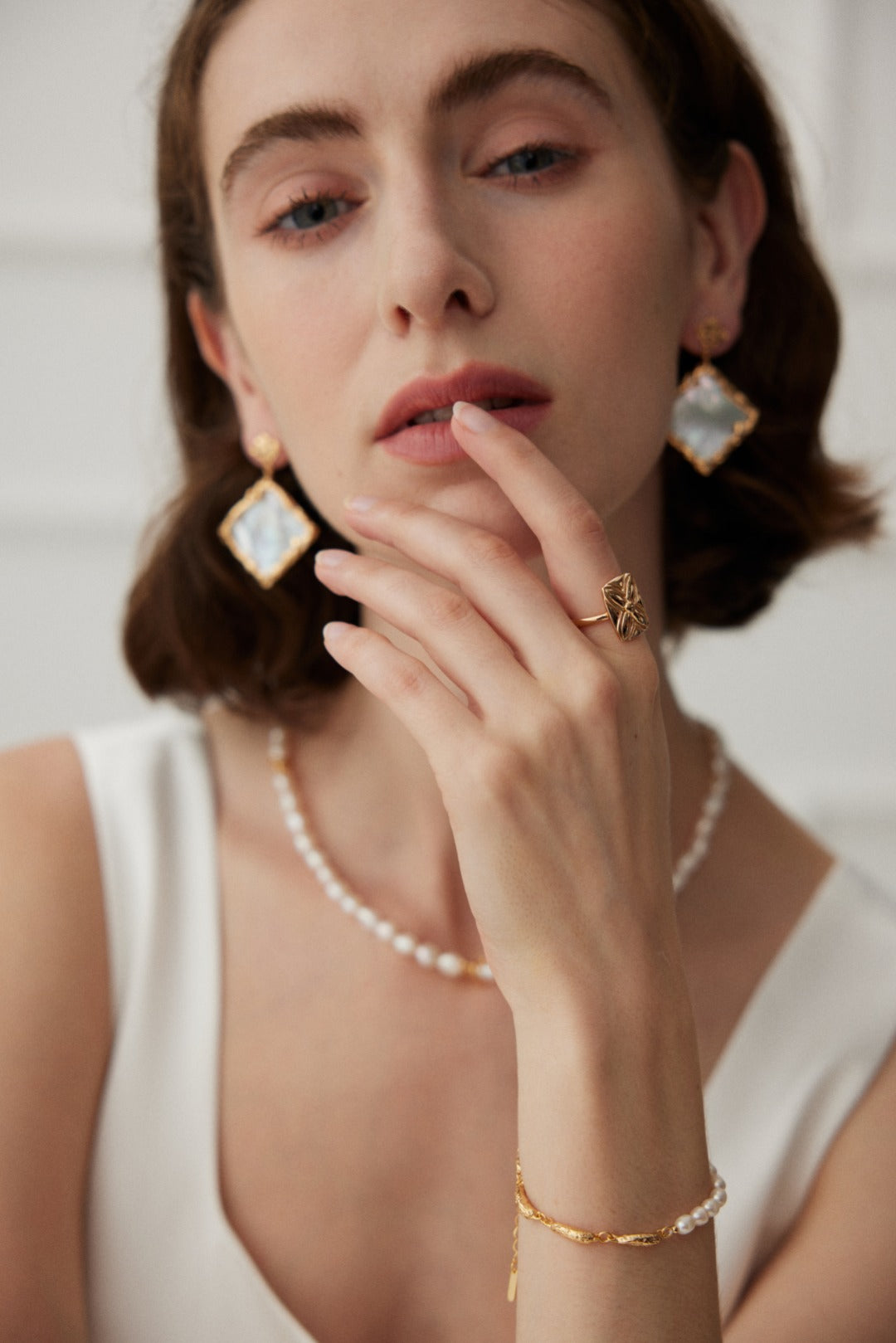 A person with shoulder-length hair is gazing thoughtfully, their hand touching their chin. They are adorned with gold and pearl jewelry, including earrings, a necklace, a bracelet, and an adjustable ring. The ring they wear is the Lucky Clover Adjustable Ring in Vintage Gold Finish - 925 Silver by Siren Mode. They are wearing a white sleeveless top against a light background.