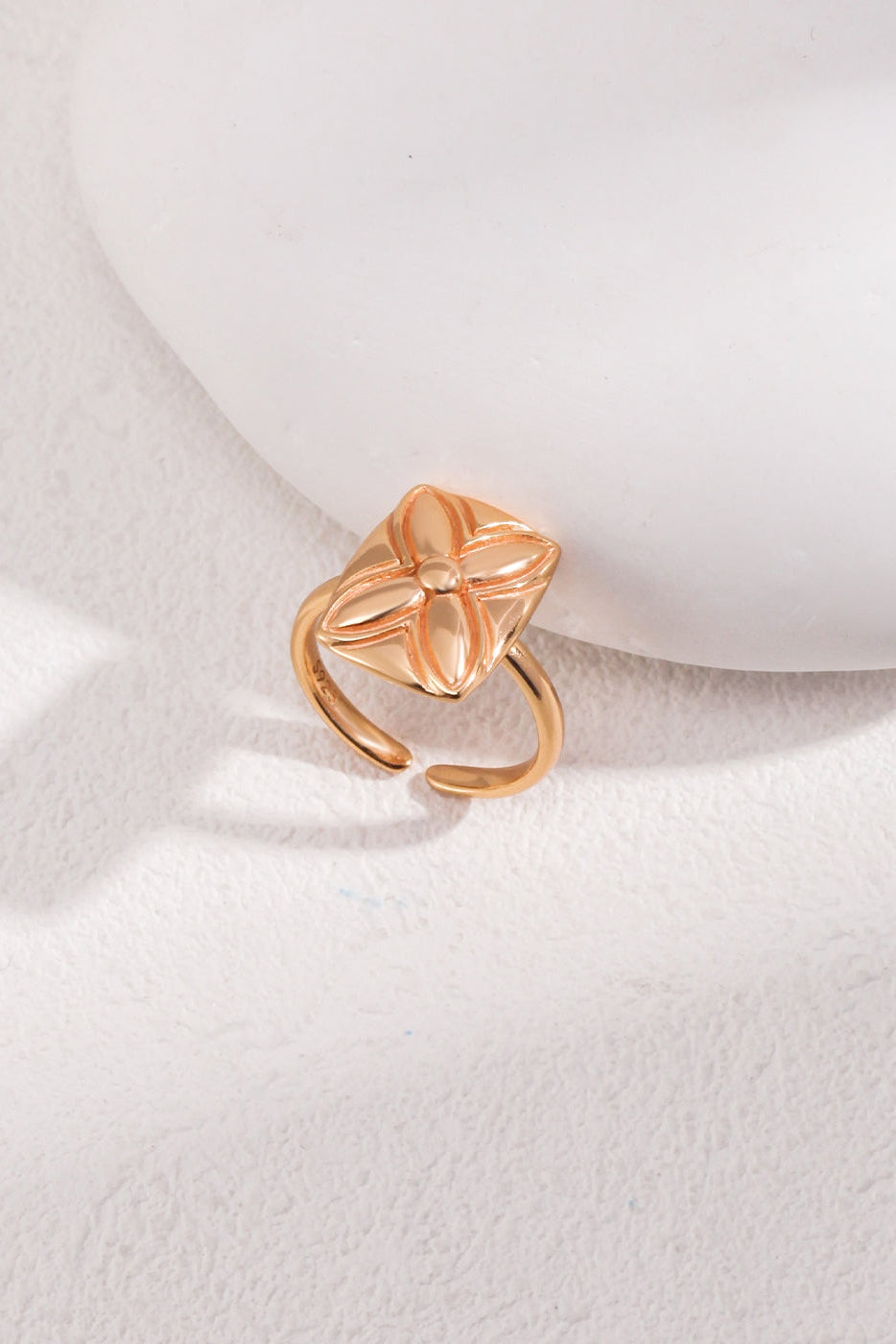 The Lucky Clover Adjustable Ring in Vintage Gold Finish - 925 Silver, by Siren Mode, is showcased against a soft white background. This exquisite ring features a unique design with a square-shaped top adorned with an intricate clover motif. The open-ended band gives it an adjustable appearance, and light and shadows enhance its details, highlighting the exceptional craftsmanship of this beautiful piece.