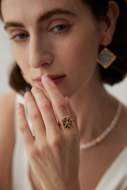 Close-up of a woman with light skin and brown hair wearing gold geometric earrings and a delicate beaded necklace. Her hand, adorned with the Siren Mode Lucky Clover Adjustable Ring in Vintage Gold Finish - 925 Silver, gently touches her face as she looks softly into the camera.