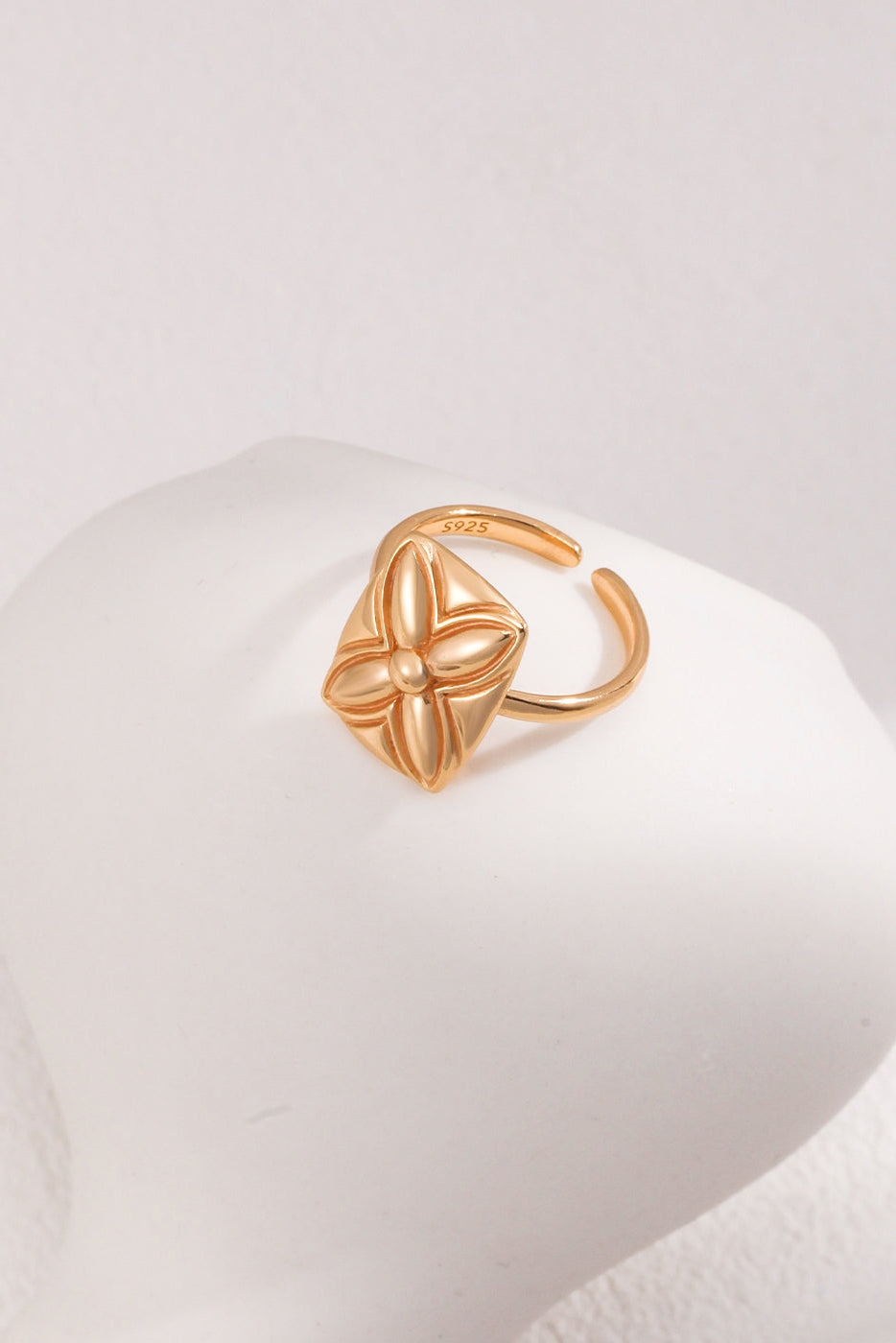 On a white mannequin hand is the Lucky Clover Adjustable Ring in Vintage Gold Finish from Siren Mode, featuring an intricate, raised floral design atop an open-ended band for customizable sizing. The soft and neutral background highlights the delicate details of this 925 silver piece with a subtle clover motif.