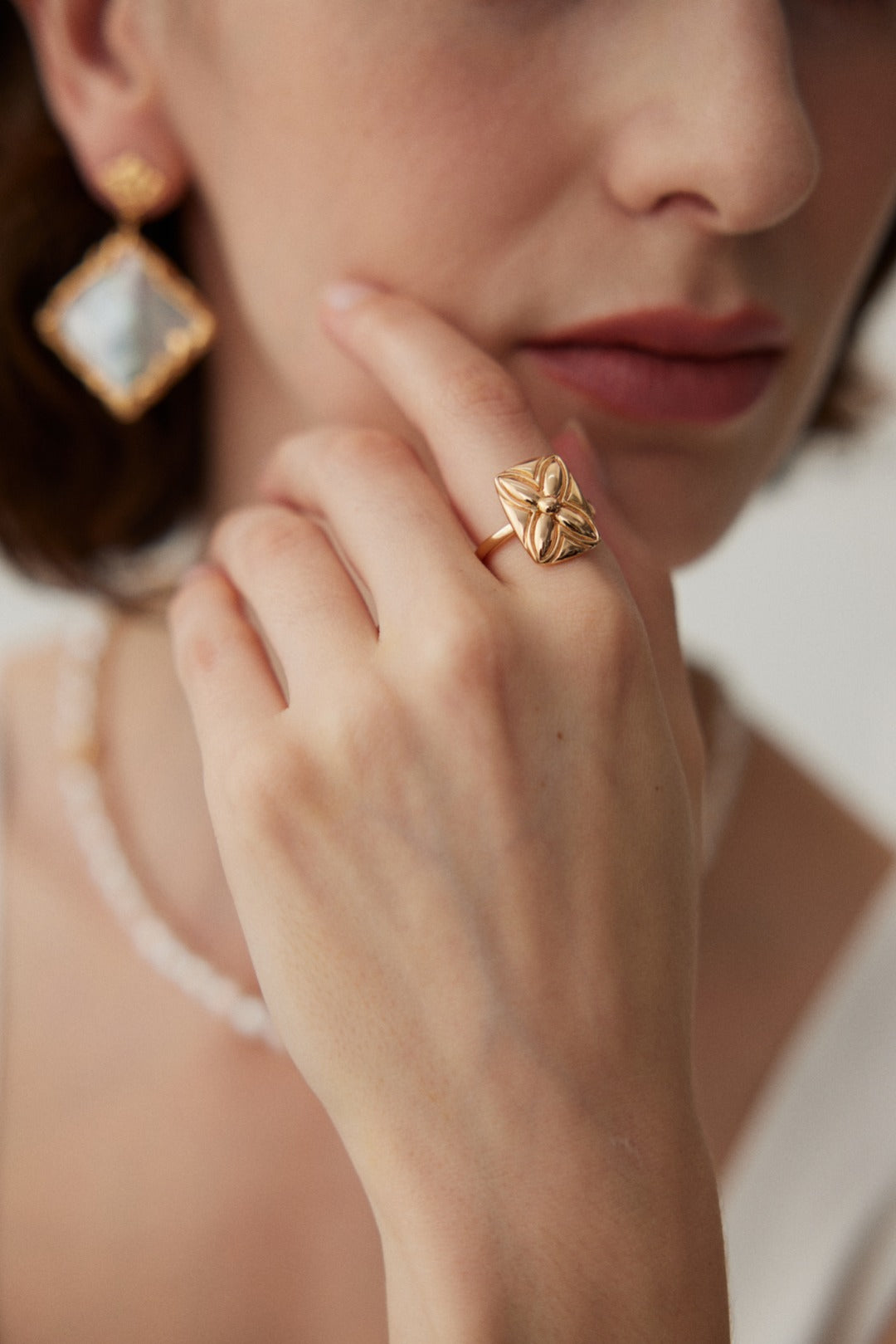 A close-up photo showcases a woman wearing a gold ring with a floral design on her finger, complemented by the Lucky Clover Adjustable Ring in Vintage Gold Finish - 925 Silver by Siren Mode. She also sports a dangling earring with a large square gemstone and a delicate necklace. Her hand gently touches her face, lips slightly parted.