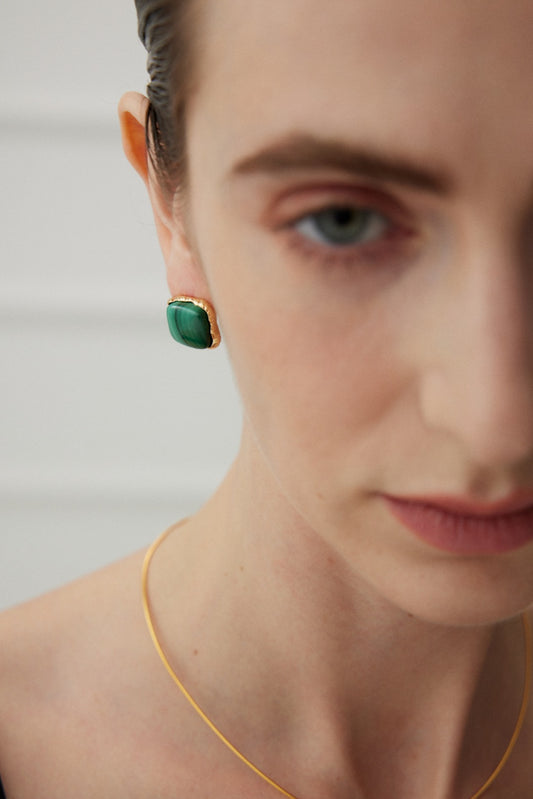 Close-up of a person with light skin and short dark hair wearing the Siren Mode Malachite Elegance Stud Earrings - Vintage Gold Frame and a delicate silver necklace. The background is softly blurred, and the person is gazing downward with a neutral expression.