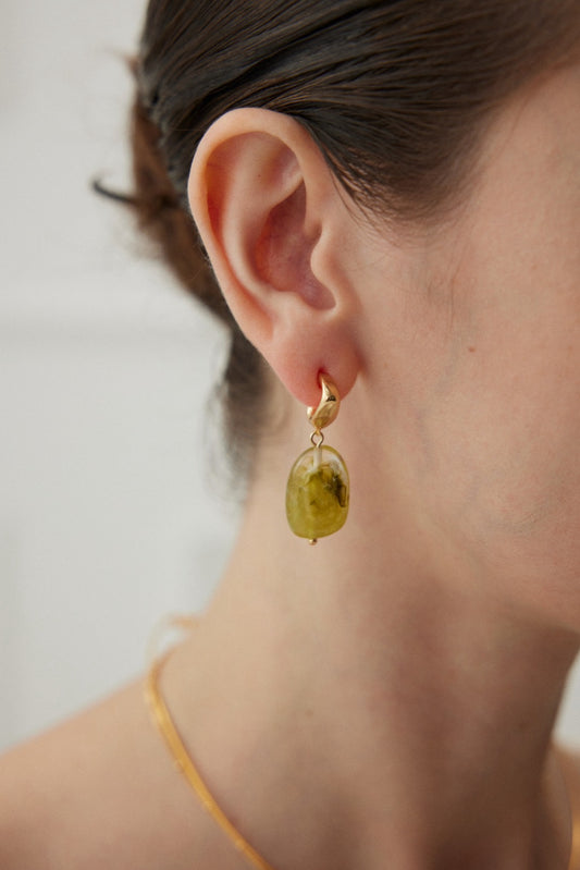 Close-up of a person's ear adorned with Siren Mode's Modern Green Resin Drop Earrings, featuring a vintage gold finish that perfectly accentuates the eco-conscious, nature-inspired fashion accessory. The person, with brown hair pulled back, also sports a thin gold necklace. The softly blurred background draws focus to these chic statement earrings.