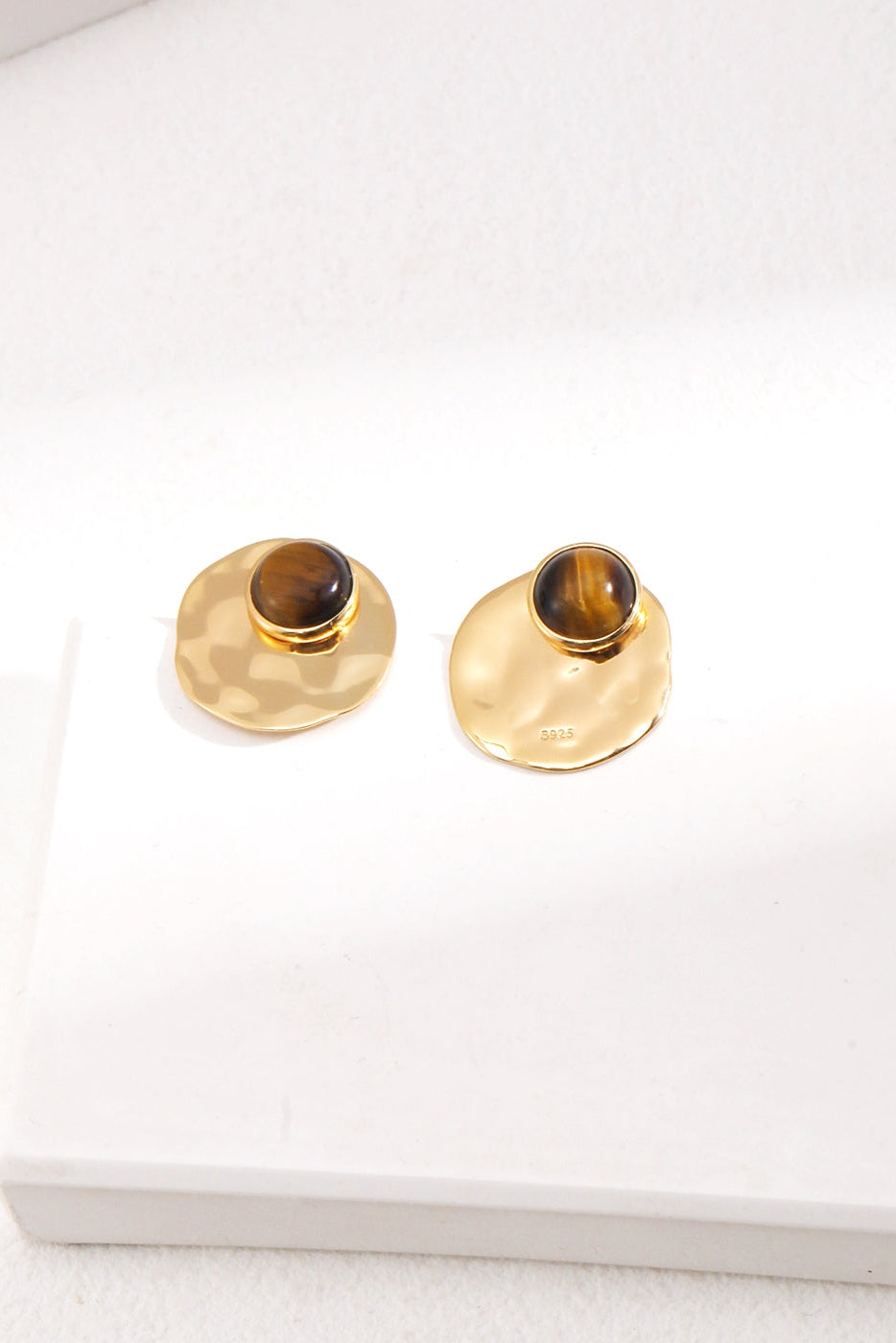 Introducing the Modern Tiger's Eye Disc Earrings - Vintage Gold Finish by Siren Mode. These earrings feature irregular, wavy gold discs with rounded, dark brown tiger's eye gemstones set near the top. They are showcased on a white surface with soft light casting delicate shadows.