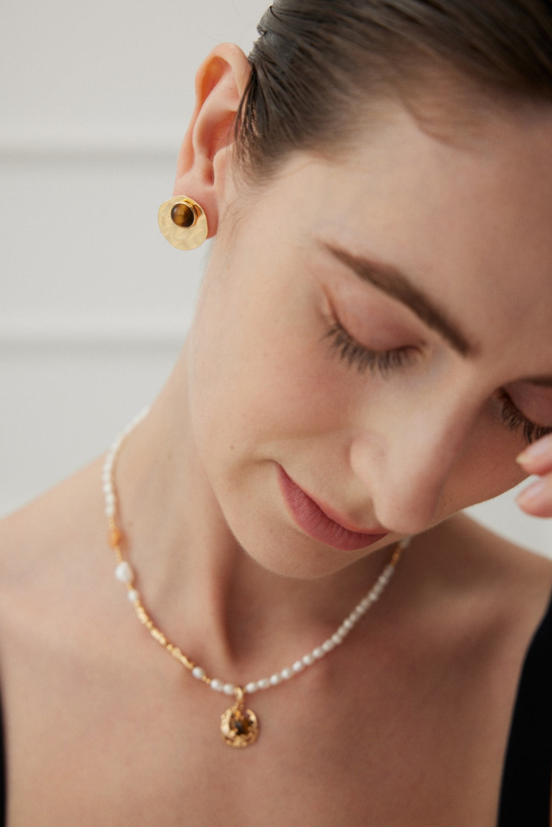 A person with short, dark hair is pictured with their eyes closed. They are wearing Modern Tiger's Eye Disc Earrings with a vintage gold finish from Siren Mode and a pearl necklace with a vintage gold pendant. Dressed in a black top, they stand against a softly blurred background in light tones, providing a delicate contrast.