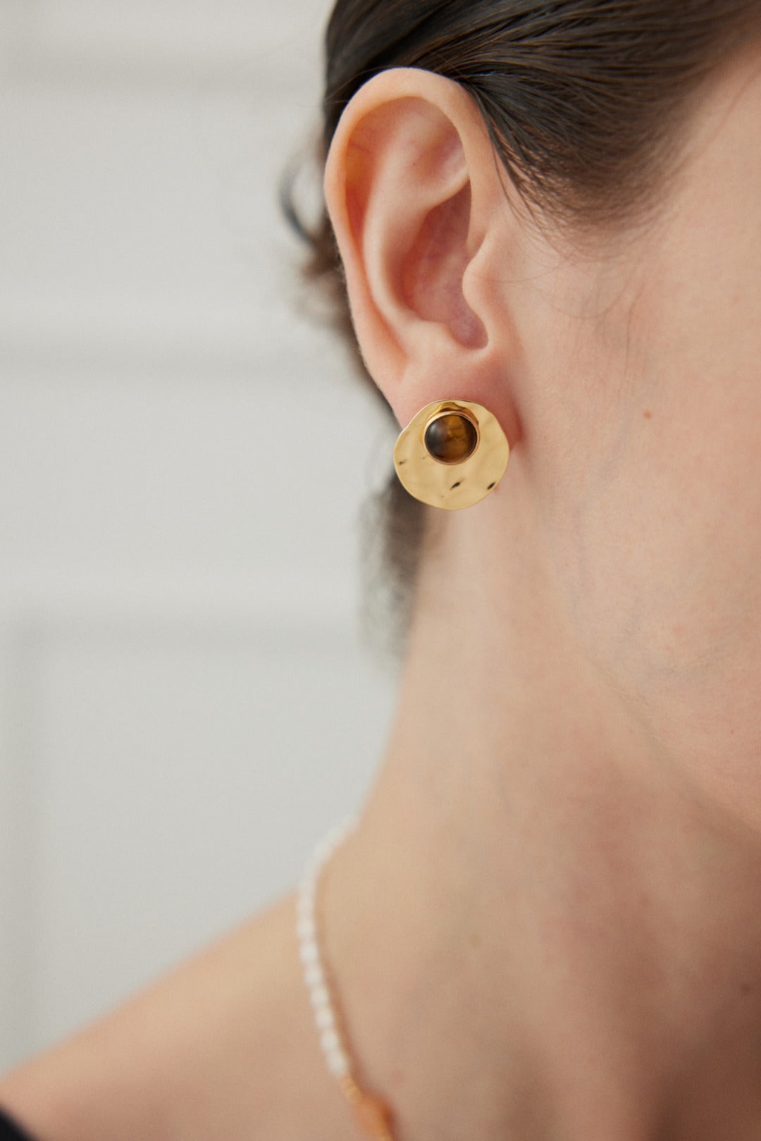 Close-up of a person wearing Siren Mode's Modern Tiger's Eye Disc Earrings in Vintage Gold Finish. The gold circular earring, featuring a brown center similar to Tiger's Eye, has a textured hammered finish. The person's ear and part of their neck are visible against a blurred, light-colored background.