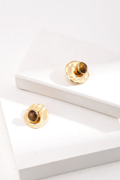 A pair of Modern Tiger's Eye Disc Earrings - Vintage Gold Finish by Siren Mode are displayed on two white rectangular blocks positioned at different heights. These stylish and elegant earrings feature a round, hammered texture with dark stone centers that catch the light to highlight their unique design.