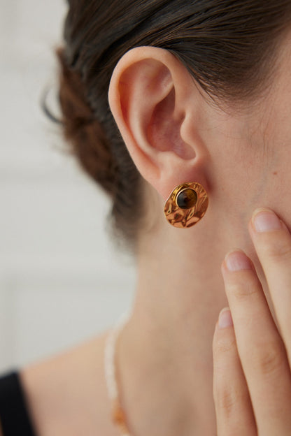 Close-up photo of someone wearing the Modern Tiger's Eye Disc Earring by Siren Mode, featuring a vintage gold finish and an intricate design with a dark tiger's eye gemstone at the center. Their hair is tied back, and they are touching their face with their fingers. The background is blurred, highlighting the stunning earring.