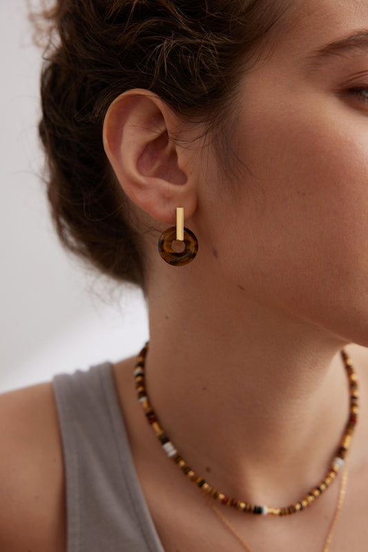 The close-up features a person with light skin wearing Siren Mode's Modern Vintage Gold Tiger's Eye Disk Earrings in 925 Silver and a beaded necklace with alternating gold, black, and white beads. They are dressed in a grey tank top, and the blurred background highlights the earthy sophistication of the jewelry.