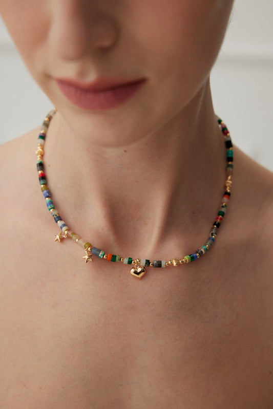 A close-up image shows a person's neck adorned with the Siren Mode Multicolored Gemstone Bead Necklace with Vintage Gold Finish. The necklace features an array of vibrant, multicolored gemstones and small vintage gold charms, including a heart and star. The beads are arranged in a mix of colors, creating an eye-catching design. The person's face is partially visible, out of focus.