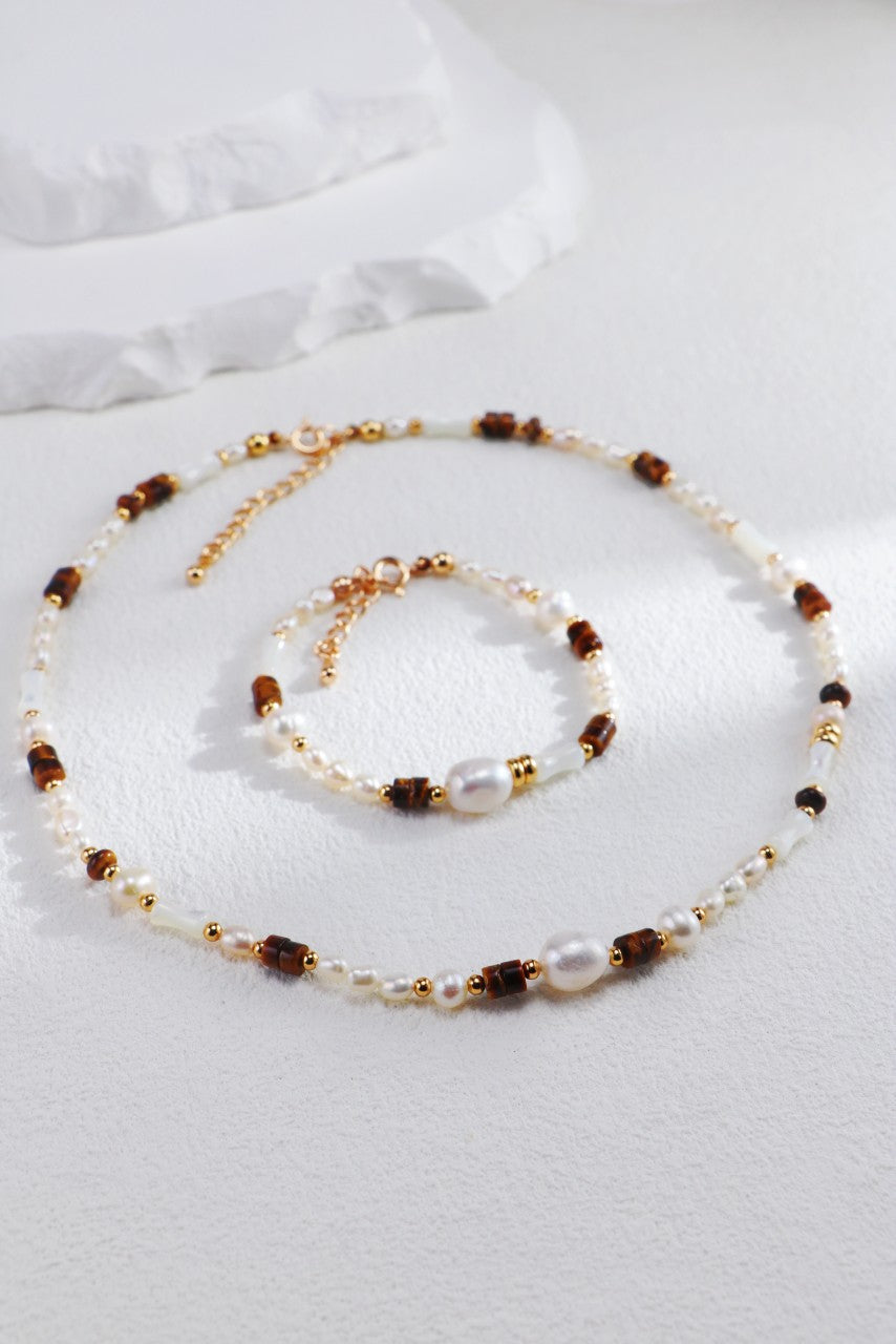 Displayed on a light surface is a set of beaded jewelry from Siren Mode, featuring a necklace and their Natural Pearl, Tiger Eye, and Mother of Pearl Bracelet. Both pieces are intricately crafted with small pearls, amber-like beads, and gold accents. Adorned with natural pearl and Tiger Eye details, the bracelet highlights focal larger pearls and an adjustable chain clasp for a perfect fit.