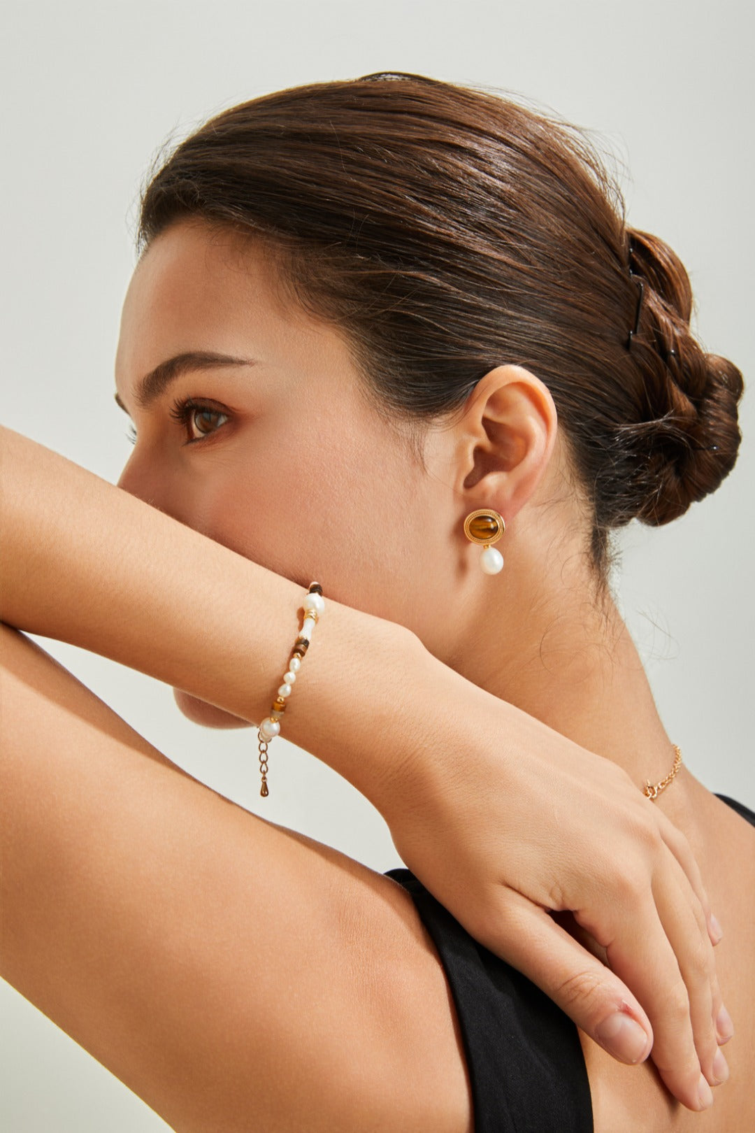 In a profile view, a person with brown hair styled in a bun rests their arm across their nose and mouth. They are adorned with a gold and natural pearl earring, a black sleeveless top, and the Siren Mode Natural Pearl, Tiger Eye, and Mother of Pearl Bracelet. The background is plain and light-colored.