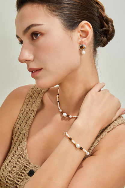 A woman with her hair in a bun wears a knitted beige top and matching jewelry, including Mother of Pearl earrings, a natural pearl necklace, and the Siren Mode Natural Pearl, Tiger Eye, and Mother of Pearl Bracelet. She gazes thoughtfully to the side, with one hand touching her neck. The background is neutral.