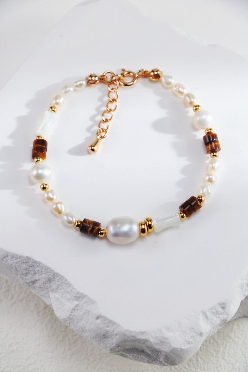 The Siren Mode Natural Pearl, Tiger Eye, and Mother of Pearl Bracelet is elegantly displayed on a white stone surface. This exquisite piece showcases a harmonious blend of white pearls, Tiger Eye beads, and subtle gold accents. For added convenience, the adjustable gold chain clasp ensures a perfect fit.