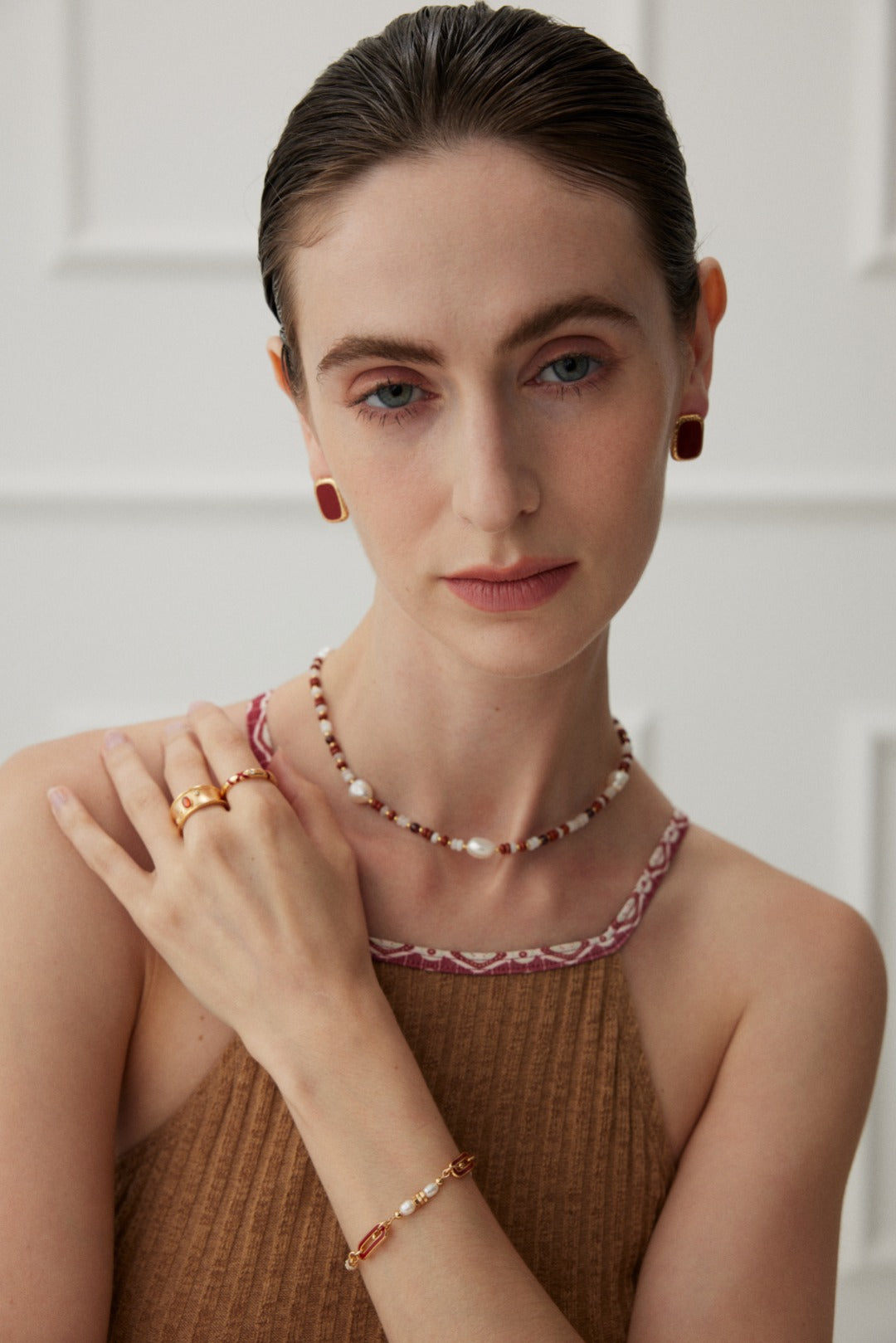 A person gazes into the camera with a calm expression, their right hand resting on their left shoulder. They are wearing a brown sleeveless top, an elegant Siren Mode Natural Pearl and Gemstone Necklace, earrings, a bracelet, and two rings. The background is softly blurred with white paneled walls.