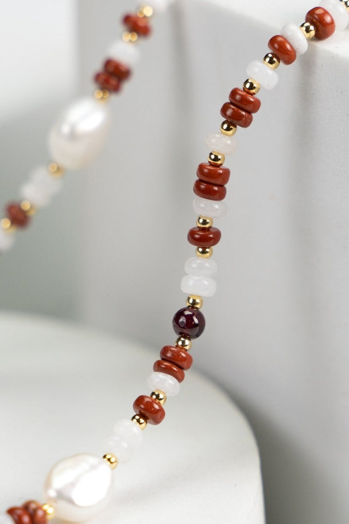 A close-up of the Siren Mode Natural Pearl and Gemstone Necklace showcases a stunning mix of small round red, white, and purple beads, accompanied by 18K gold accents and uniquely shaped white pearls. The necklace is artistically draped over a white geometric backdrop.