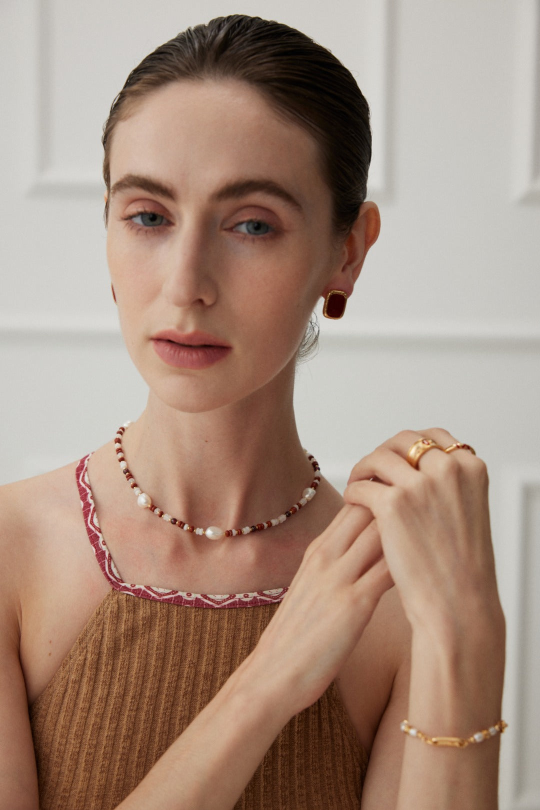 A person with a neutral expression is wearing a sleeveless brown top featuring red trim. Their dark hair is slicked back, and they are accessorized with Siren Mode's Natural Pearl and Gemstone Necklace in 18K gold, alongside gold and red earrings, and multiple gold rings. The backdrop consists of a white paneled wall.