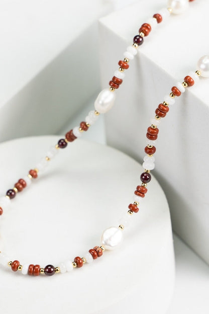 A delicate beaded necklace from Siren Mode, the Natural Pearl and Gemstone Necklace, features a combination of natural pearls, small red coral beads, and tiny gold accents artistically arranged. This exquisite piece is elegantly draped over a white geometric display, showcasing its intricate and colorful design.