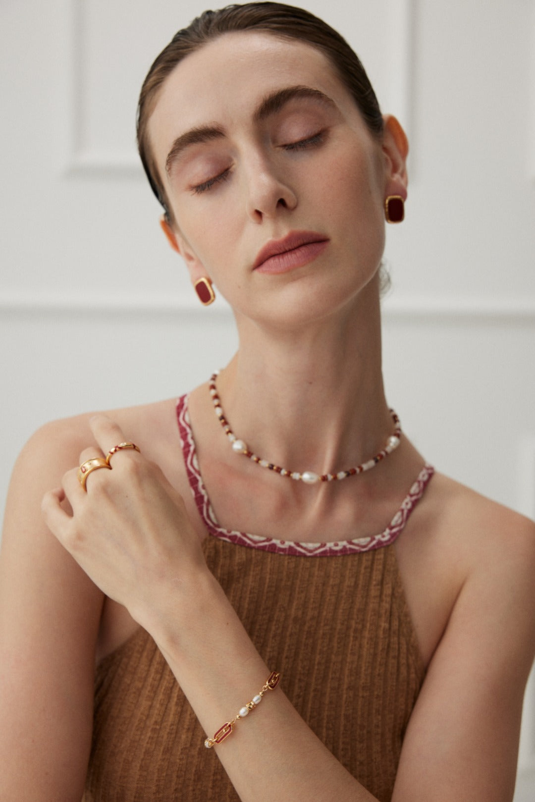 A woman with light skin and dark blond hair is pictured with her eyes closed, wearing red earrings, a Natural Pearl and Gemstone Necklace by Siren Mode, a bracelet with red and gold details, and a brown sleeveless top with red and white trim. She is also wearing two rings on her left hand.