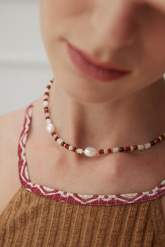 Close-up of a person wearing a Siren Mode Natural Pearl and Gemstone Necklace, featuring a mix of small beads and larger pearls. The top they are wearing is made of a brown ribbed fabric with a patterned trim in red and white. The image focuses on the necklace and the neckline.