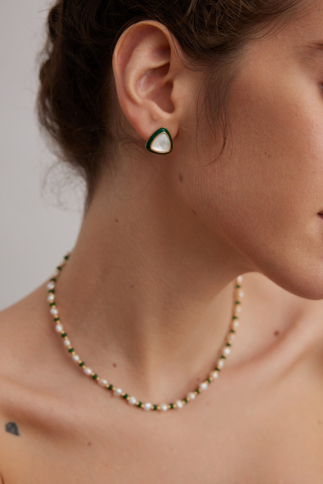 A close-up shot of a person wearing the Siren Mode Natural Pearl and Green Gemstone Necklace and a pair of triangle-shaped earrings. The focus is on the jewelry, with the background blurred. The person's face is partially visible, highlighting their neck and ear.