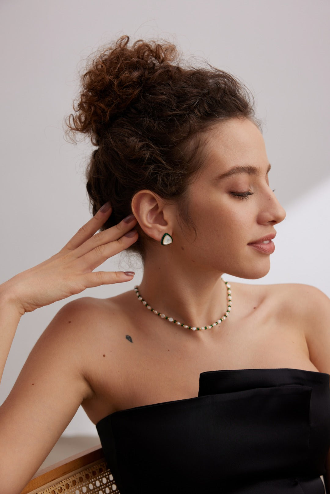 A woman with curly hair in a bun is sitting and posing with her head turned to the side. She is wearing a strapless black dress and a Siren Mode Natural Pearl and Green Gemstone Necklace, complemented by matching pearl earrings. One hand is raised near her face, revealing a small heart tattoo on her shoulder.