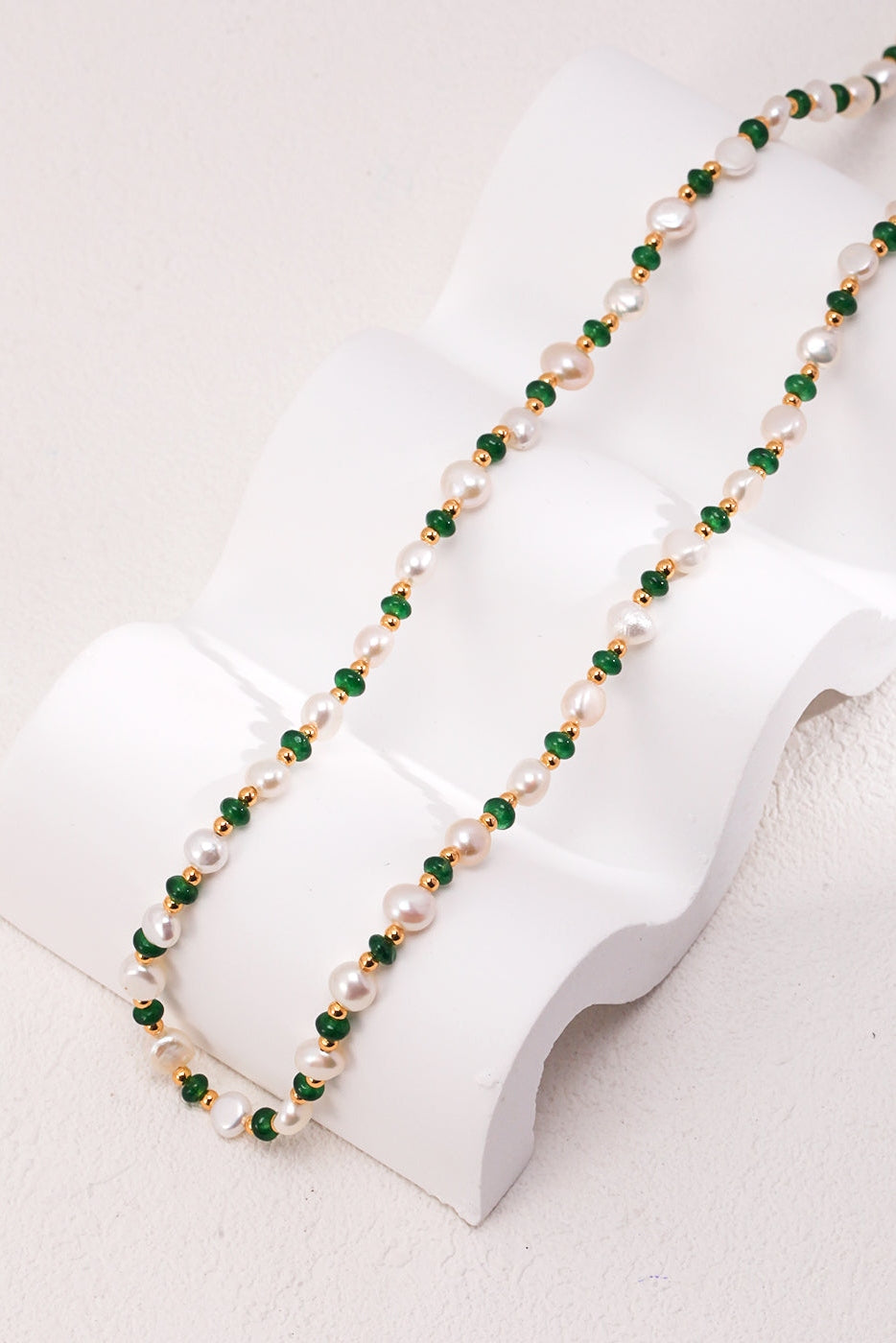 The Siren Mode Natural Pearl and Green Gemstone Necklace is gracefully displayed on a sculpted white stand, draped elegantly to highlight its exquisite design. This necklace features alternating white pearls and small green beads, beautifully accented with vintage gold elements.