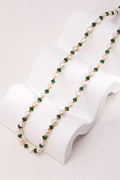 The Siren Mode Natural Pearl and Green Gemstone Necklace is gracefully displayed on a sculpted white stand, draped elegantly to highlight its exquisite design. This necklace features alternating white pearls and small green beads, beautifully accented with vintage gold elements.