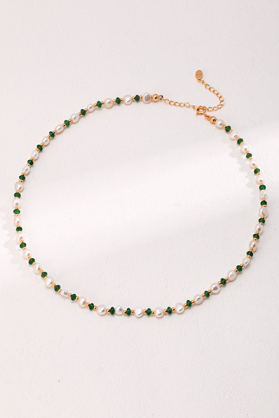 The Natural Pearl and Green Gemstone Necklace by Siren Mode is elegantly displayed on a light background. This delicate necklace features a pattern of small green and white beads with vintage gold accents and includes an adjustable clasp. The light casts soft shadows across the beautiful piece.