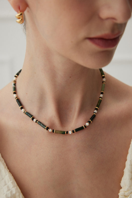 Close-up image of a woman's neck wearing the Siren Mode Nature's Elegance Necklace - Vintage Gold with Agate and Onyx, adorned with green and white cylindrical beads, and black and white round beads. She has small gold hoop earrings and is dressed in a light-colored, textured garment. The background is softly blurred.