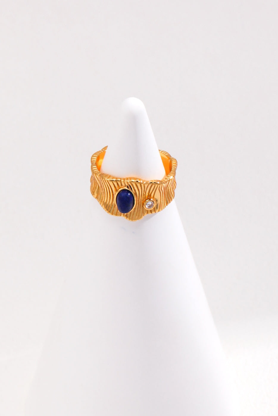 The Regal Vintage Gold Lapis Lazuli Ring in 925 Silver by Siren Mode features a textured surface and a round Lapis Lazuli gemstone set alongside a small, clear gem. This adjustable ring is showcased on a white, cone-shaped stand against a plain white background.