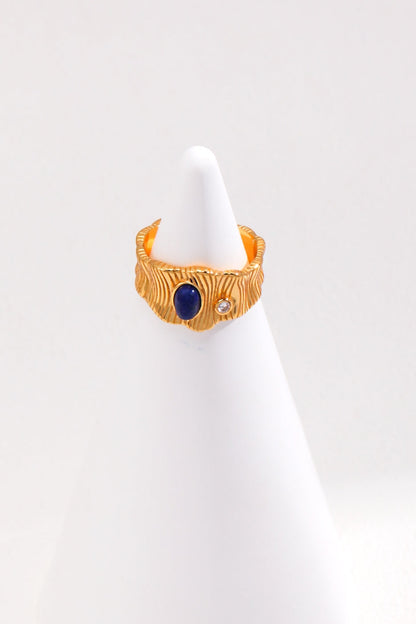 The Regal Vintage Gold Lapis Lazuli Ring in 925 Silver by Siren Mode features a textured surface and a round Lapis Lazuli gemstone set alongside a small, clear gem. This adjustable ring is showcased on a white, cone-shaped stand against a plain white background.