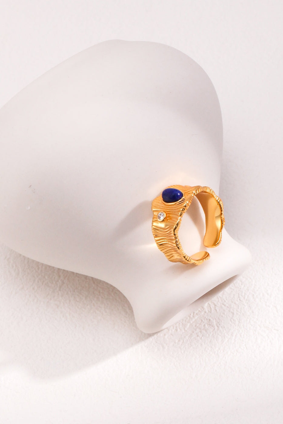 The Regal Vintage Gold Lapis Lazuli Ring in 925 Silver by Siren Mode, displaying an intricate design with a small blue stone and a tiny clear gem, is elegantly set against a white surface with a soft-focus background. The adjustable ring rests gracefully on the curved side of a white matte vase, adding subtle contrast to the composition.