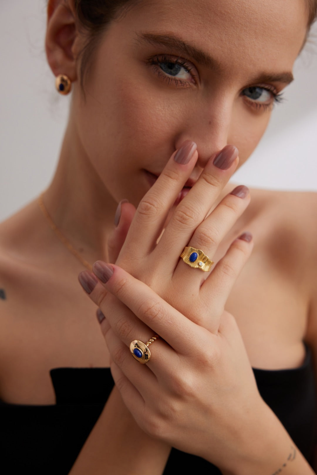 A person with light skin and painted nails, wearing a black strapless top, poses with hands raised near their face. They adorn themselves with matching gold jewelry featuring Siren Mode's Regal Vintage Gold Lapis Lazuli Ring in 925 Silver, including earrings, rings, and a necklace.