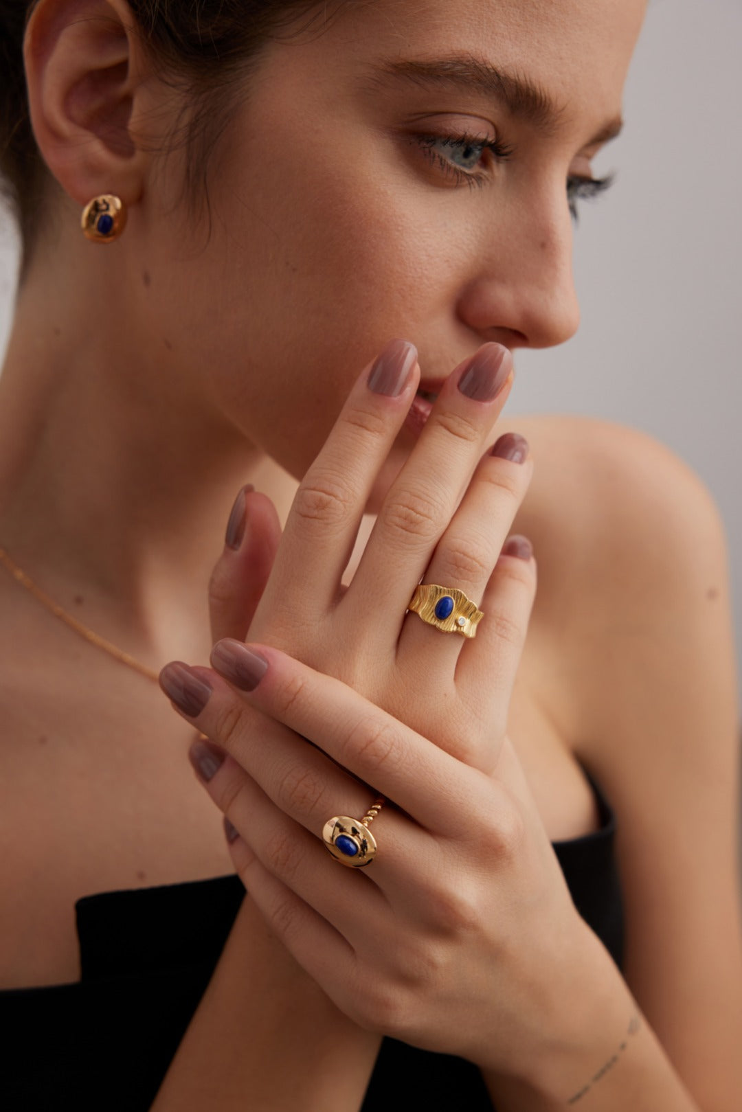 A woman with neatly styled hair and natural makeup poses, displaying jewelry. She wears a vintage gold ring with blue stones, along with matching gold earrings and a necklace. Her hand, showcasing an elegant Regal Vintage Gold Lapis Lazuli Ring in 925 Silver by Siren Mode, is raised to her face against a neutral background, highlighting her accessories and nails.