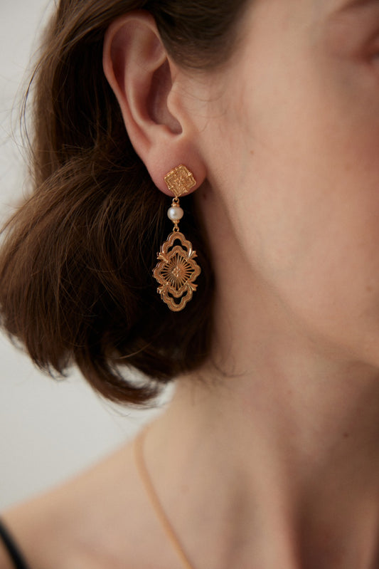 Close-up of a woman's ear adorned with Siren Mode's Regl Vintage Gold Pearl Drop Earrings with Intricate Design in 925 Silver. The earrings feature a decorative top with a small, detailed square and a larger, ornate dangling piece, showcasing natural pearl drops in between. The neutral background and her brown hair frame the elegant design.