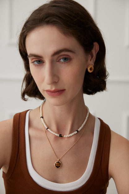 A woman with short brown hair wears a sleeveless brown and white top. She accessorizes with vintage gold jewelry, including Siren Mode's Royal Tiger's Eye and Pearl Vintage Gold Earrings, a necklace featuring a gold pendant, and a beaded choker-style necklace. The background is softly blurred, keeping the focus on her.