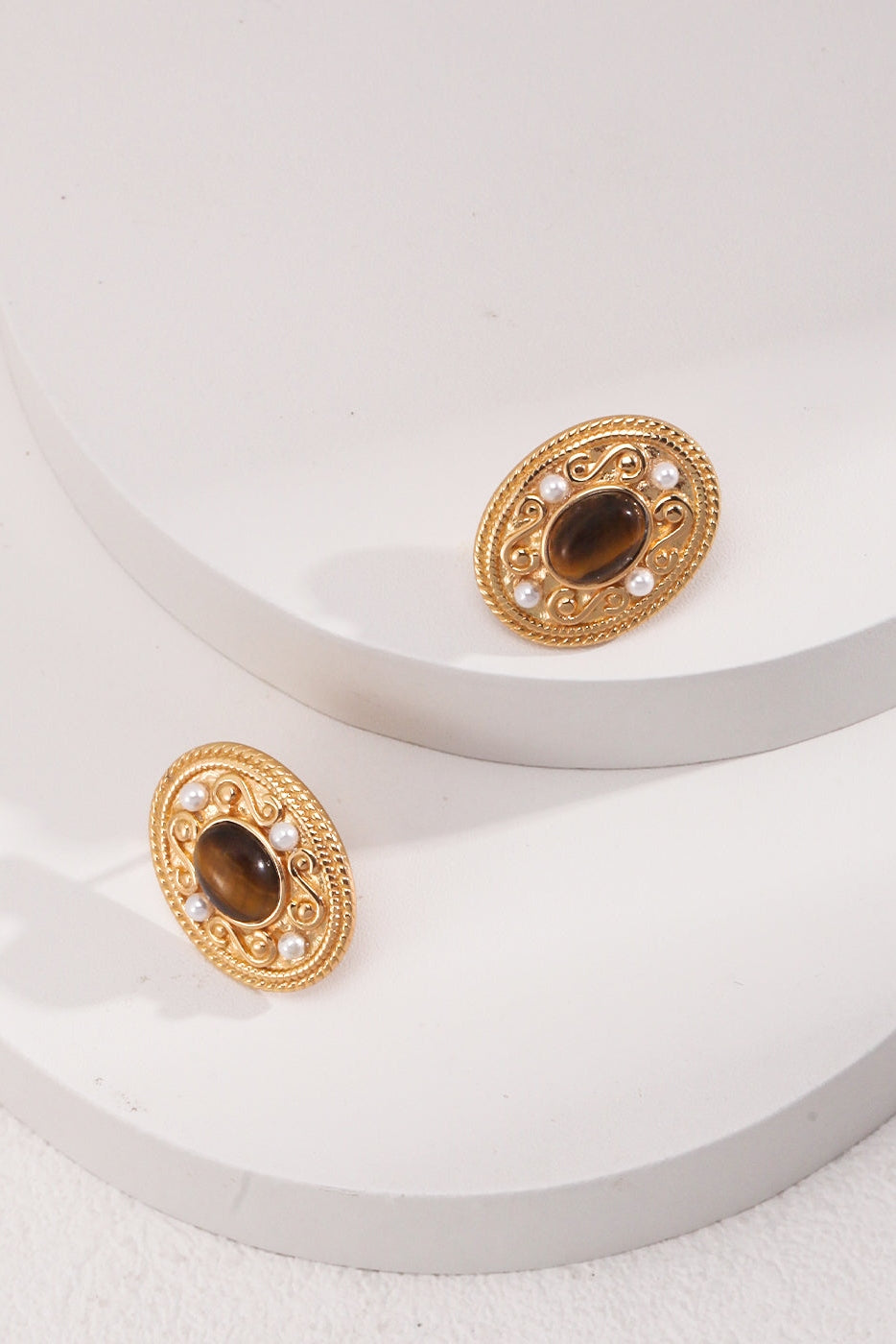 A pair of Siren Mode's Royal Tiger's Eye and Pearl Vintage Gold Earrings, elegantly displayed on two white circular platforms. Each earring showcases an intricate filigree design with a dark central Tiger's Eye gemstone, surrounded by small natural pearls. The background features a smooth, matte surface that enhances the vintage charm of these exquisite gold earrings.