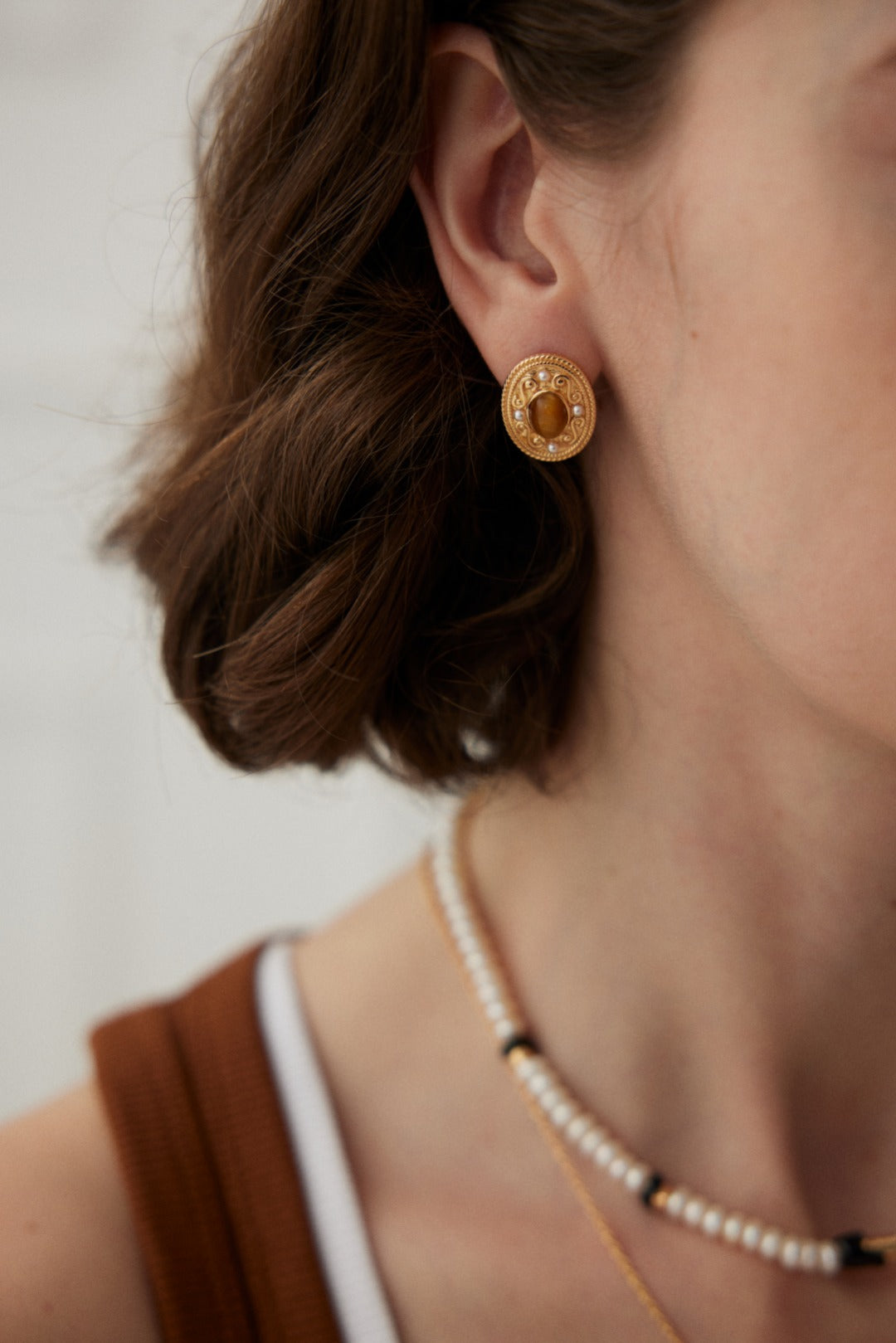 Close-up of a person wearing vintage gold jewelry—a pair of Royal Tiger's Eye and Pearl Vintage Gold Earrings by Siren Mode, featuring a circular design, paired with a matching necklace. Their short brown hair is styled in loose waves, and they are wearing a rust-colored tank top. The background is softly blurred.