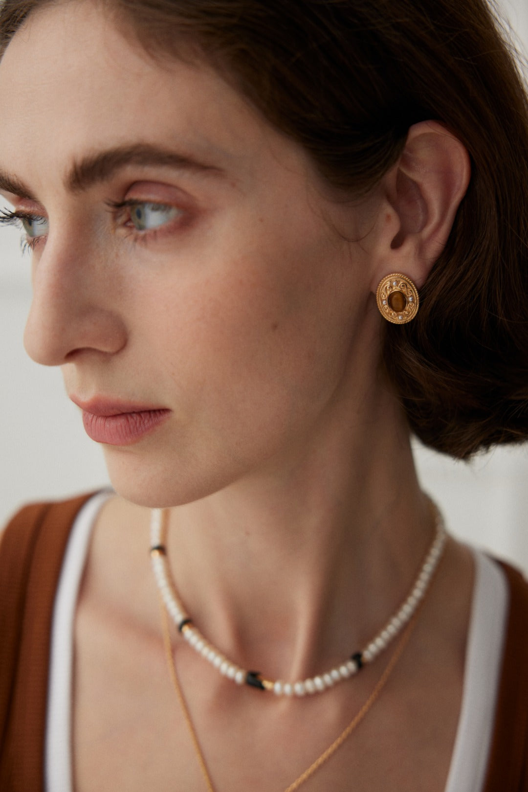 A woman with light skin and brown eyes looks to the side. She has shoulder-length brown hair and wears vintage gold jewelry, including the "Royal Tiger's Eye and Pearl Vintage Gold Earrings" by Siren Mode, along with two necklaces: one with natural pearls and black beads, and the other with a small gold pendant. She is dressed in a white top under a brown cardigan.