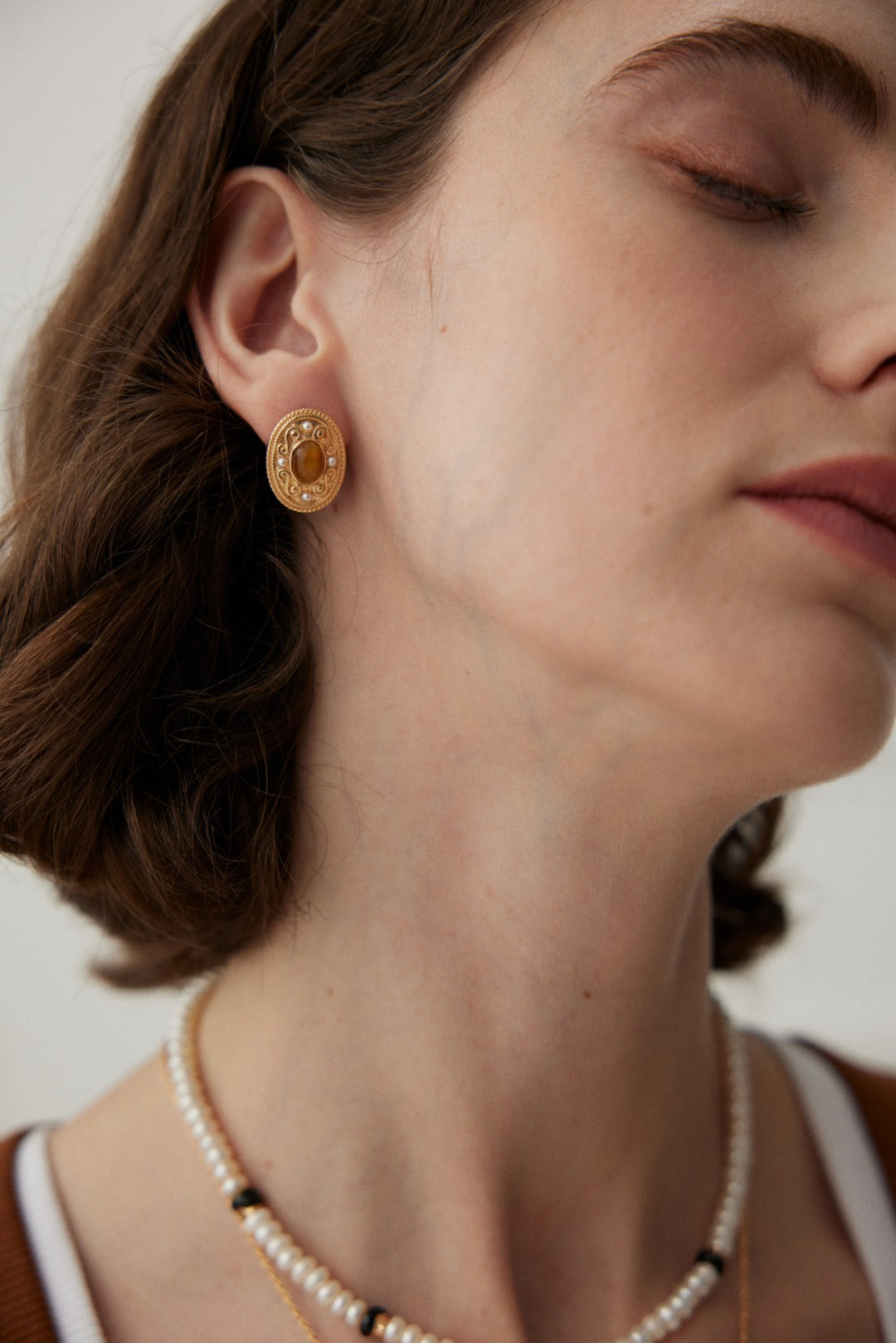 Close-up of a person with short, dark hair wearing the Royal Tiger's Eye and Pearl Vintage Gold Earrings from Siren Mode. They wear a white pearl necklace with black accents and have their eyes closed, facing slightly to the side. They are dressed in a brown and white top.