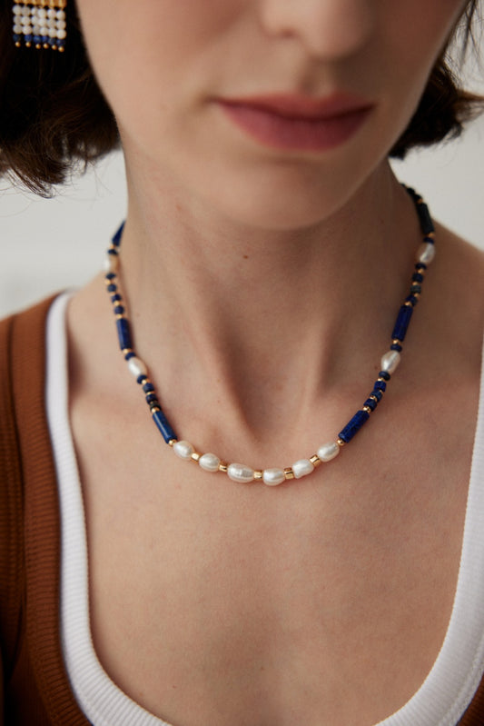 A close-up of a person wearing an elegant Siren Mode accessory: the Sophisticated Lapis Lazuli and Pearl Necklace in Vintage Gold. The person has short brown hair and is dressed in a white shirt with a brown outer layer.