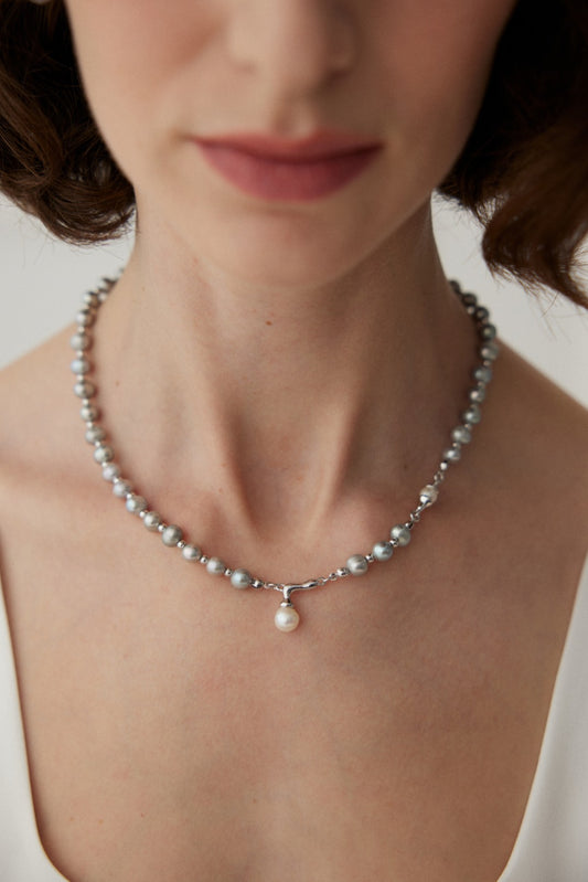 A close-up image of a person wearing the Sophisticated White Gold Pearl Necklace with Adjustable Chain in 925 Silver by Siren Mode. The necklace features small, natural pearls and a single larger pearl pendant at the center. The person is wearing a white top and has a light complexion, while the background is blurred.