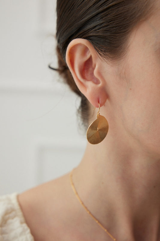 Close-up image of a person wearing a large, round Spiral Radiance Earring in Vintage Gold Finish by Siren Mode, featuring a textured pattern and revealing part of their ear and neck. They are also wearing a delicate gold chain necklace. The background is blurred and neutral-colored.