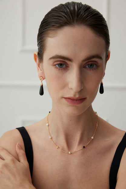 A woman with slicked-back hair wears a black top, Starry Night Blue Sandstone Drop Earrings - Vintage Gold by Siren Mode, and a delicate gold necklace with pearls and small beads. She looks forward with a neutral expression. The background features a softly focused white wall with some molding details.