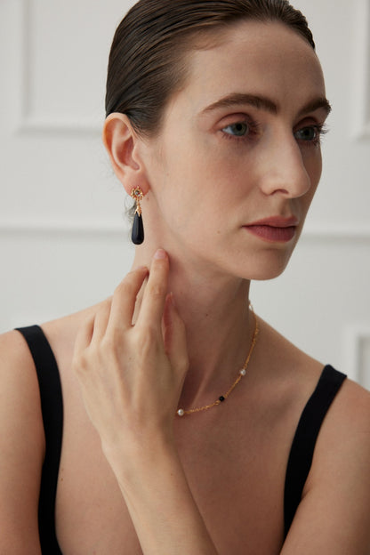 A woman with light skin and slicked-back hair is shown wearing a black sleeveless top. She is touching her ear, adorned with Siren Mode's Starry Night Blue Sandstone Drop Earrings in vintage gold. A delicate gold necklace with small beads graces her neck, and the background remains plain white.