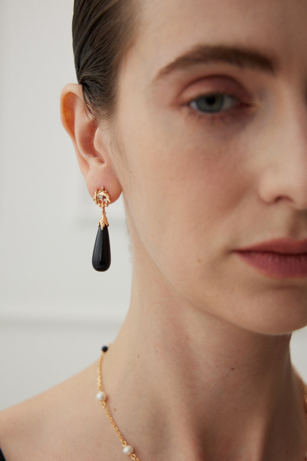 Close-up of a person's face focusing on their earring. The earring is the "Starry Night Blue Sandstone Drop Earrings - Vintage Gold" by Siren Mode, featuring a black teardrop-shaped pendant that exudes vintage charm. The person has a subtle makeup look and wears a necklace adorned with small pearls. The background is blurred, emphasizing the earring and part of the face.