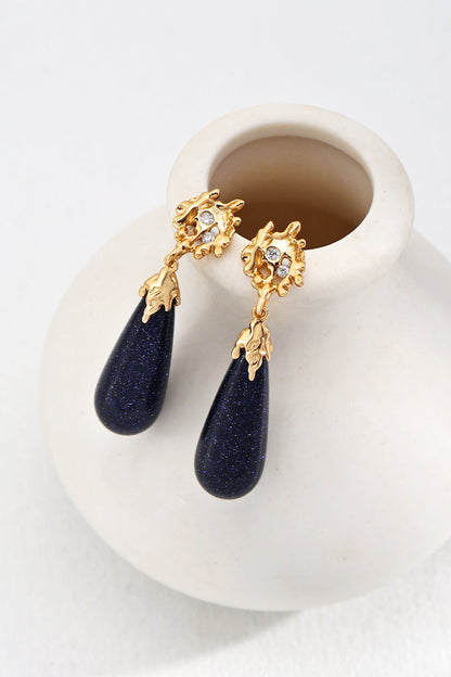 The Starry Night Blue Sandstone Drop Earrings - Vintage Gold by Siren Mode feature elegant golden floral tops adorned with small clear gemstones. The dark navy blue teardrop-shaped pendants, crafted from blue sandstone, add a vintage charm as they rest gracefully on the rim of a white matte vase.